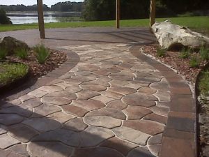 Flagstone-pattern paver walkway curving past a boulder and planting bed, water beyond