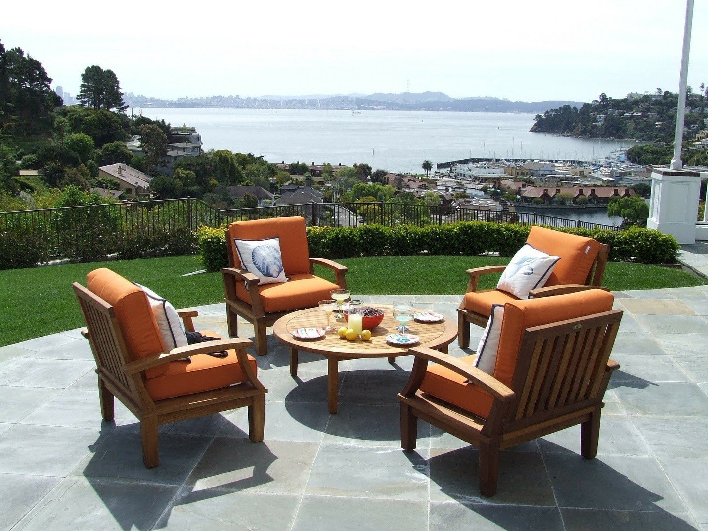 Two orange-cushioned armchairs and a coffee table on a stone patio overlooking a bay