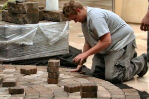 Worker kneeling to lay tan paver blocks on a bed of sand