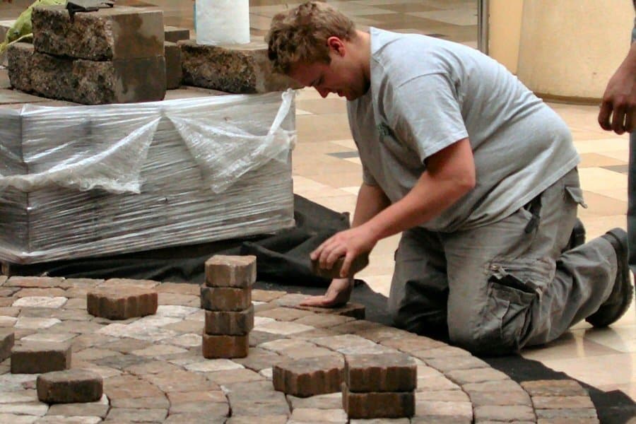 Worker kneeling to lay tan paver blocks on a bed of sand