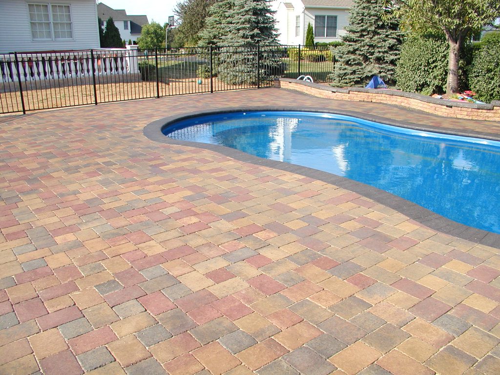 Kidney shaped pool surrounded by sealed red and tan pavers, white fence behind