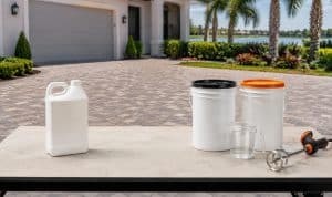 A single-component sealer jug beside a two-part sealer kit, measuring cup and mixing paddle on a paver driveway